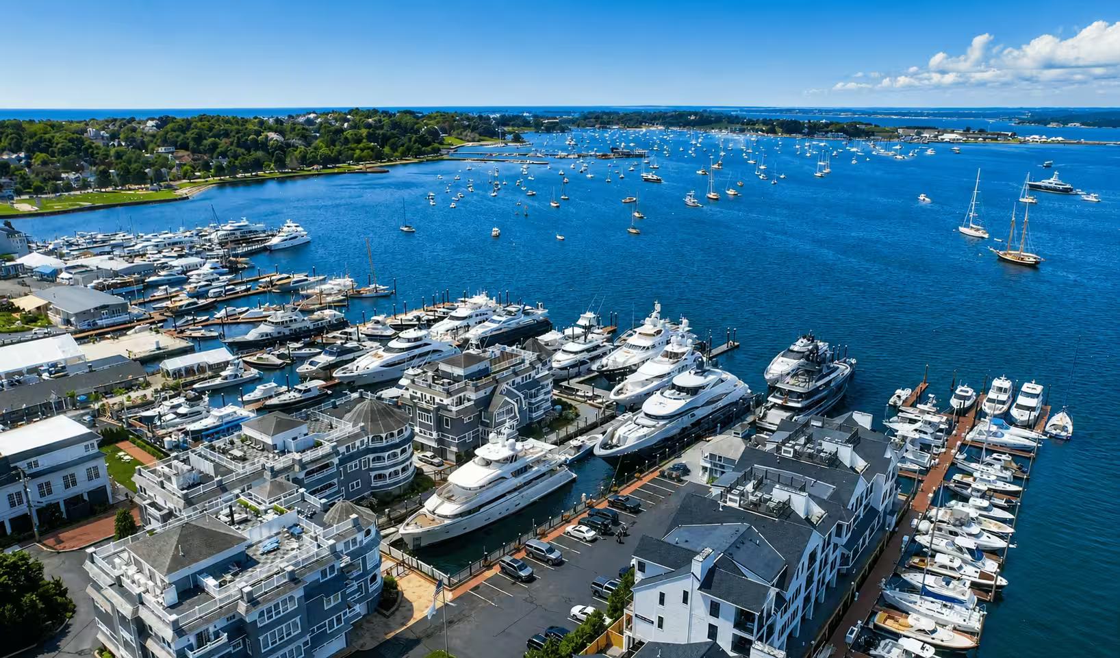 Aerial view of a busy marina — yachts and boats docked along the piers of a waterfront town, open ocean beyond.