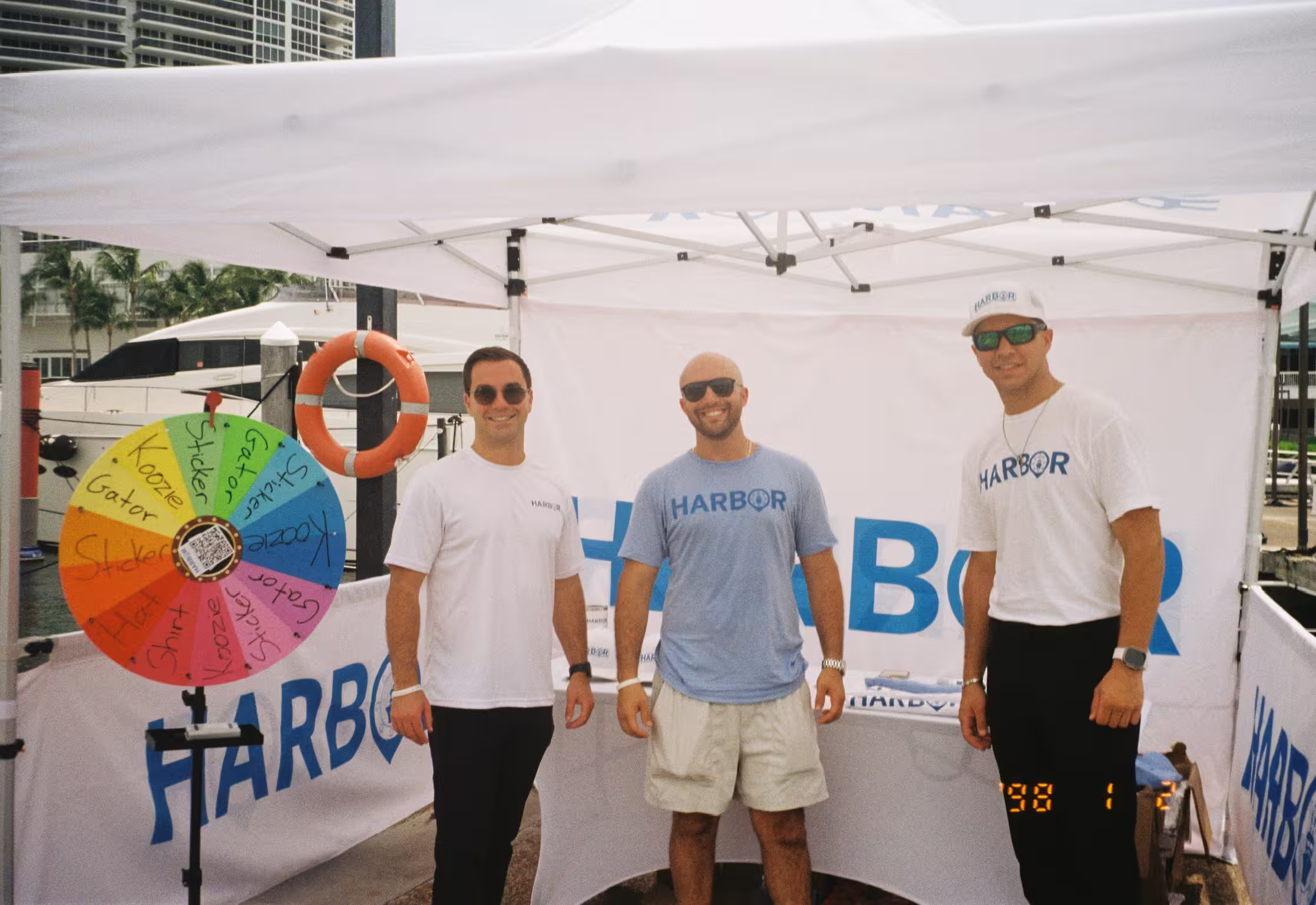 Three members of the Harbor team in branded shirts at a marina event booth, a prize wheel and Harbor banners beside them and a yacht docked behind.