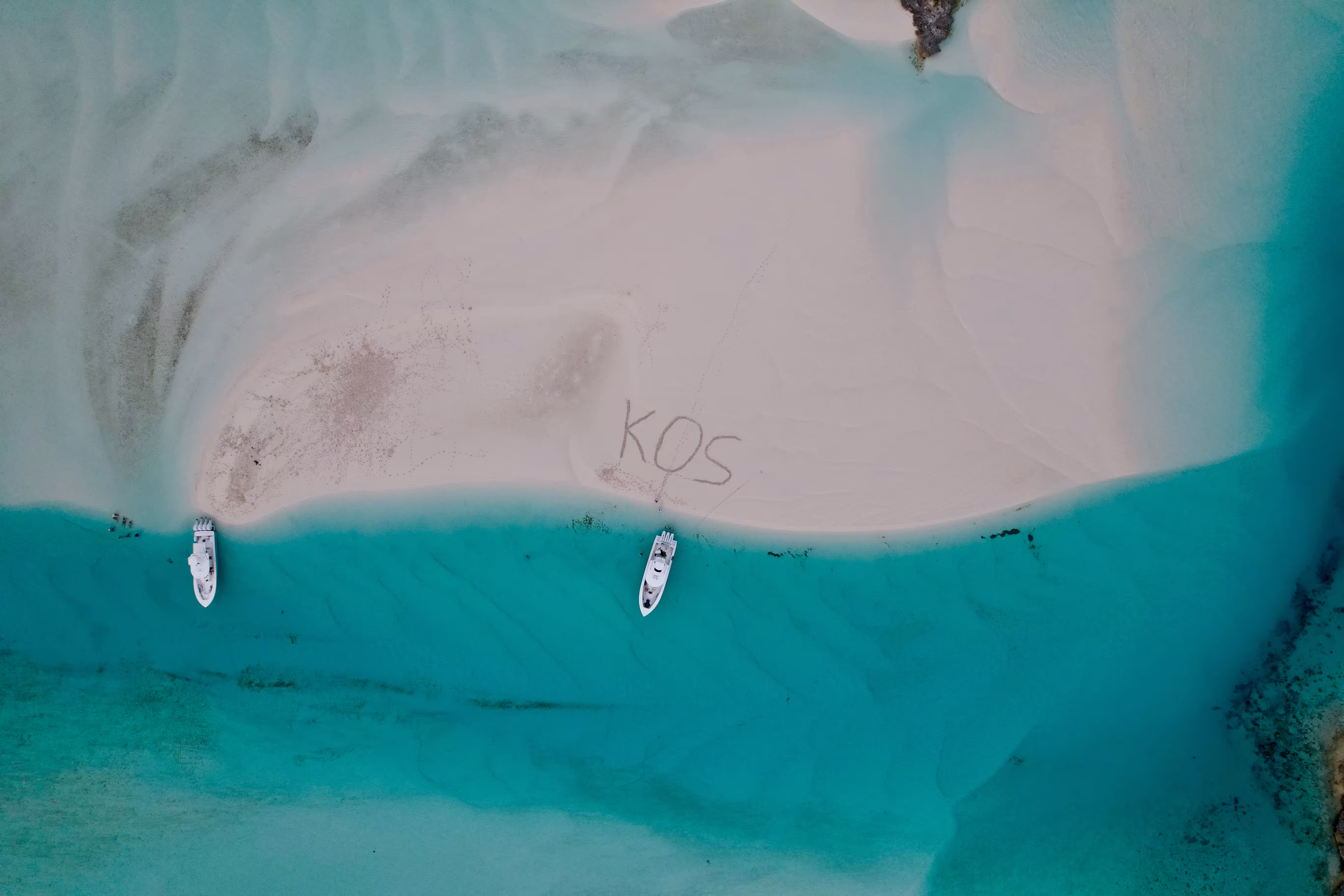 Aerial view of two boats moored at a white sandbar in turquoise Bahamas water, with "KOS" drawn in the sand.