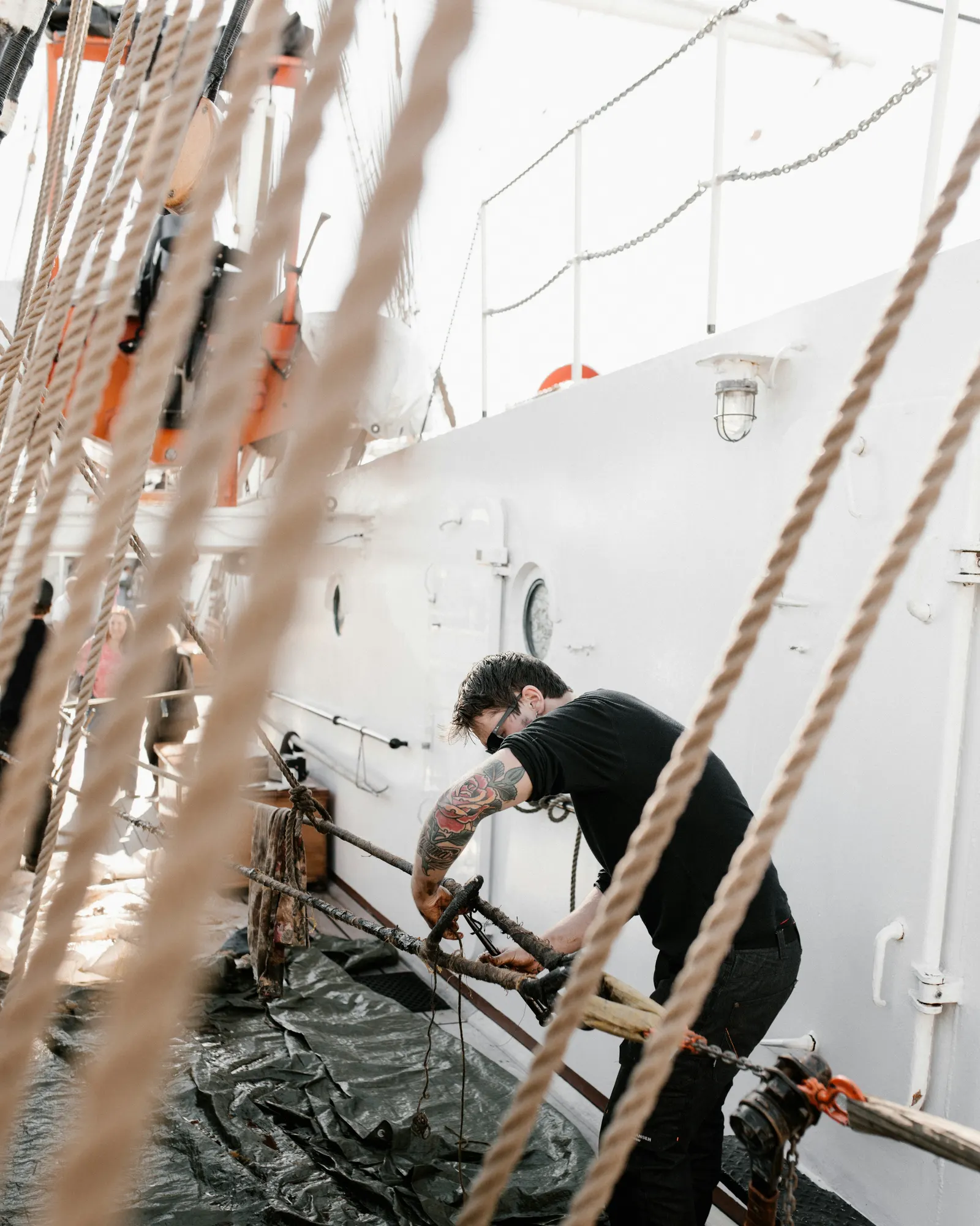 A crew member working on deck hardware aboard a vessel, rigging in the foreground.